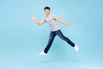Fototapeta na wymiar Jumping shot of ecstatic young Asian man with arms raising in mid air isolated in blue studio background