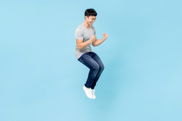 Fototapeta na wymiar Portrait of smiling young happy Asian man clenching fists and jumping on isolated light blue studio background