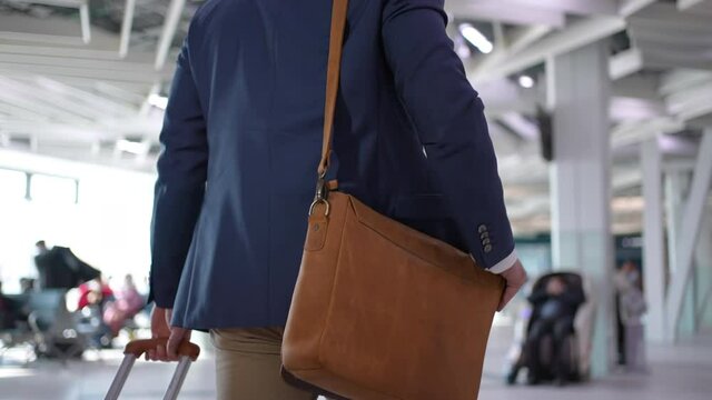Businessman passenger pulling suitcase, handbag luggage in modern airport terminal. Young traveler man carries shoulder bag walking in airport, close-up rear view, Travel business concept