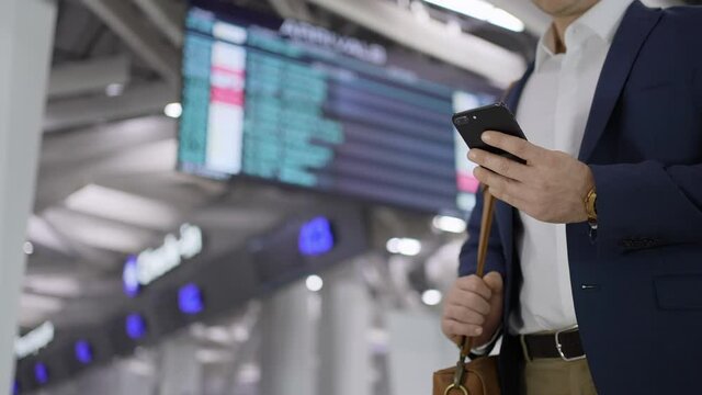 Hand of tourist traveler man using mobile phone of plane arrivals board timetable in airport terminal for travel. Businessman typing text message on smartphon check-in at flight, Technologies business