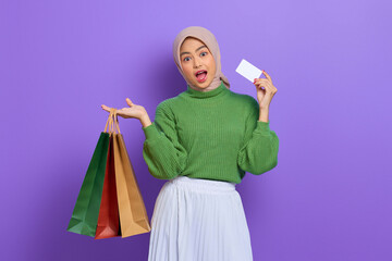 Shocked beautiful Asian woman in green sweater holding shopping bags and credit card isolated over purple background