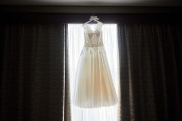 Beautiful beige embroidered wedding dress hanging on hanger against window in the room, copy space, close up. Bridal morning preparations