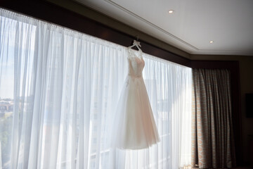 Beautiful beige embroidered wedding dress hanging on hanger against window in the room, copy space, close up. Bridal morning preparations