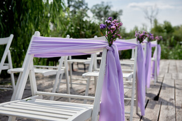 White wedding chairs with fresh flowers and purple cloth on each side of archway outdoods, copy space.  Empty wooden chairs prepared for wedding ceremony
