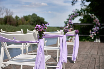 White wedding chairs with fresh flowers and purple cloth on each side of archway outdoods, copy space.  Empty wooden chairs prepared for wedding ceremony