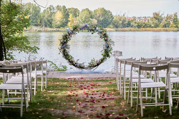 Place for wedding ceremony on river beach outdoors, copy space. Circle wedding arch decorated with flowers and chairs on each side of archway. Wedding setting