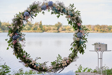 Circle wedding arch decorated white flowers and greenery by lake outdoors, copy space. Wedding setting. Floral composition
