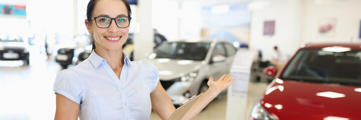 Woman salesman with clipboard in her hands showing new car in dealership