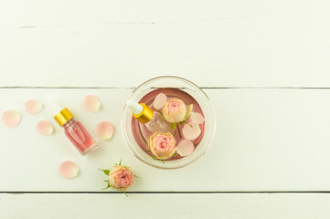 glass bowl with rose water and rose petals, two bottles of cosmetic rose oil on a white wooden background. top view.