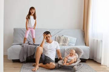 Indoor shot of exhausted attractive young adult man sitting on floor near sofa and keeping hand on his head, being tired of noisy little kids, wearing white t shirt and jeans short.
