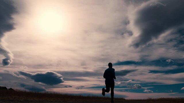 Striking black silhouette outline of a male jogger, jogging against a dramatic sky.  The jogger runs away from the camera in a wide angle from left to right.  The sun flares around the outline.