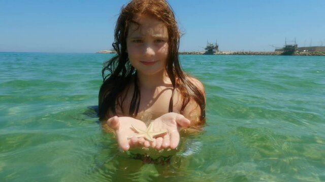 Beautiful adorable redhead child girl bathing in seawater in Italy while holding starfish in hands with trabocchi in background. Slow-motion