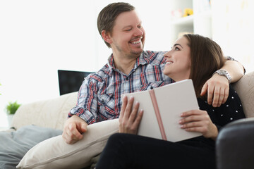 Middle aged father and young daughter reading book together sitting on couch at home