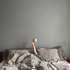 Hand's of young woman with coffee mug in bed with neutral grey bed linen cloth. Minimal happy...