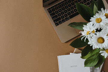 Aesthetic minimalist home office workspace desk. Laptop computer, notebook, daisy flowers bouquet on dark brown background. Flat lay, top view work, business concept