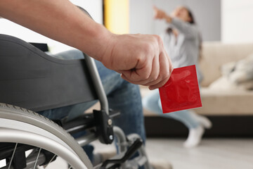 Man sit in wheelchair hold package of condom, woman laugh on couch pointing with finger