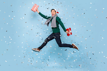 Full body portrait of jumping young Asian man with Christmas gift box in isolated light blue studio...