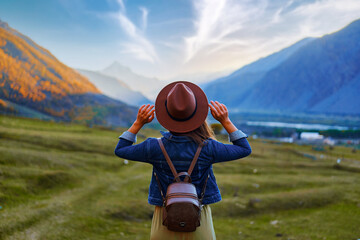 Back view of hipster girl wanderer wearing hat and backpack traveling alone on mountain valley in...