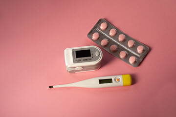 Pulse oximeter for measuring level of oxygen in blood, thermometer, tablets in blister,  on a pink background. Health device without people.
