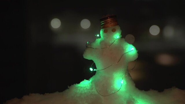 From left to right pan of little snowman litted by LED with pea eyes and bottle metal cap on head standing on snowdrift when night and snowing. Background is dark, out of focus with bokeh