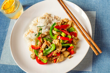 Piece of chicken meat with red and green pepper, chopstick on plate, glass of water with orange on blue.