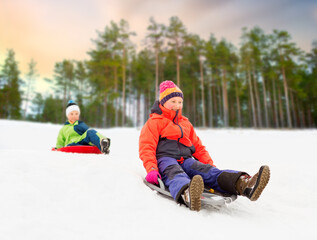Fototapeta na wymiar childhood, sledging and season concept - happy little kids sliding on sleds down snow hill in winter over snowy park or forest background