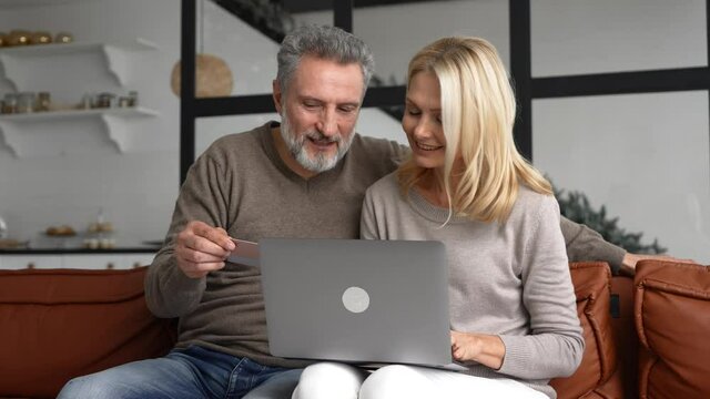 Cheerful middle-aged couple watching at laptop screen and checking credit card number to make online transaction, smiling mature spouses are shopping online, transferring money, online banking