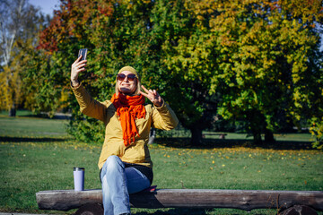 Cheerful girl in bright clothes and sunglasses sitting on park bench and taking selfie on smartphone against autumn trees. Attractive woman makes hand gestures on the background of nature. Copy space.