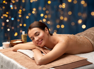 wellness, beauty and relaxation concept - beautiful young woman lying with exfoliating sea salt scrub on skin of her back at spa over christmas lights on dark blue background