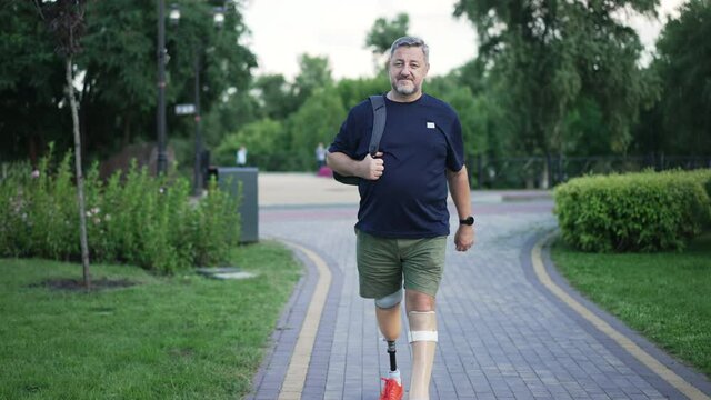 Front view portrait confident grey-haired male amputee walking in slow motion in summer park looking around. Wide shot positive Caucasian man with grey hair and prosthetic legs strolling outdoors