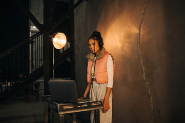 Beautiful female DJ plays a music set at the event, listens to the musician in headphones and looks at the laptop screen.