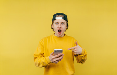 Shocked guy in yellow sweatshirt isolated on yellow background with smartphone in hands, looking at camera and pointing finger at smartphone
