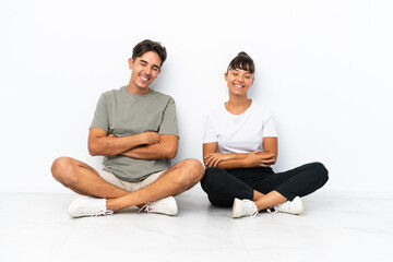 Fototapeta na wymiar Young mixed race couple sitting on the floor isolated on white background keeping the arms crossed while smiling