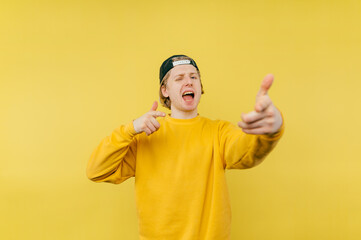 Positive guy in a cap shows his fingers to the camera and winks with a smile on his face on a yellow background, wearing a yellow sweatshirt