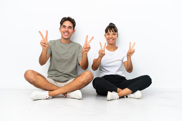 Fototapeta na wymiar Young mixed race couple sitting on the floor isolated on white background smiling and showing victory sign with both hands
