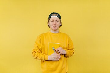 Positive student guy with notebook and pen in hands isolated on yellow background, looking at camera with smile on face.