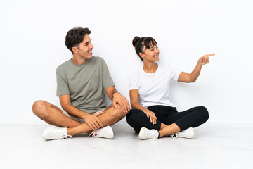 Fototapeta na wymiar Young mixed race couple sitting on the floor isolated on white background presenting an idea while looking smiling towards