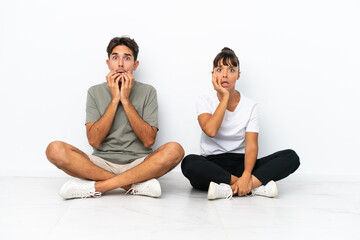 Fototapeta na wymiar Young mixed race couple sitting on the floor isolated on white background surprised and shocked while looking right