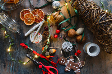 Making home made garlad from natural materials - dry orange chips, ginger bread cookies, arcons, ribbons, cinnamon sticks and pine cones, eco friendly, on wooden table with lights, flat lay