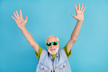 Photo of pretty friendly retired man wear vintage jeans waistcoat dark glasses rising arms smiling isolated blue color background