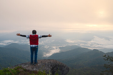 Asian man with freedom arms open standing on rock ledge of mountain peak at Pha Tang view point 104 with river view and sea mist and sun rise in heavy fog sky. travel place in Chaing Rai, Thailand