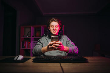 A positive guy gamer in a headset around his neck sits at night in a room at the computer and uses a smartphone with a serious face in purple light.