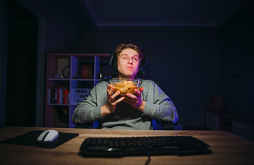 Funny guy gamer in a headset sits at a table at night with a plate of chips in his hands and eats with pleasure in the cross between the game.