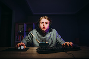 Funny gamer plays games with a focused face and eats noodles in the bedroom at home, looking at the camera. The gamer has dinner with Chinese food © bodnarphoto