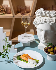 Grilled salmon with asparagus and sauce on a white plate on a blue background. Nearby olives in a white plate and a glass of white wine