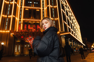 Stylish lady in a black jacket in winter poses on the background of architecture decorated with light for the holidays, looking at the camera with a serious face.