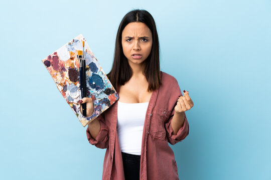 Young artist woman holding a palette over isolated blue background making money gesture but is ruined.