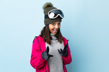 Mixed race skier girl with snowboarding glasses over isolated blue background surprised and pointing front.