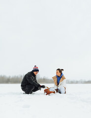 Fototapeta na wymiar Couple playing with a cute dog in a jacket in the snow on a winter cold day. Walking with a dog Jack Russell and playing in the snow.