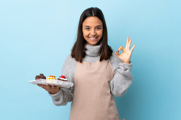 Pastry chef holding a big cake over isolated blue background showing ok sign with fingers.